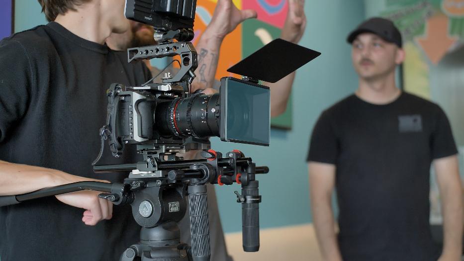 Behind-the-scenes image of Black Gate Productions crew during a commercial video shoot. Features a Lumix cinema camera rig on a tripod with matte box, monitor, and follow focus system, as a videographer gestures mid-discussion and another team member observes in a creative studio setting